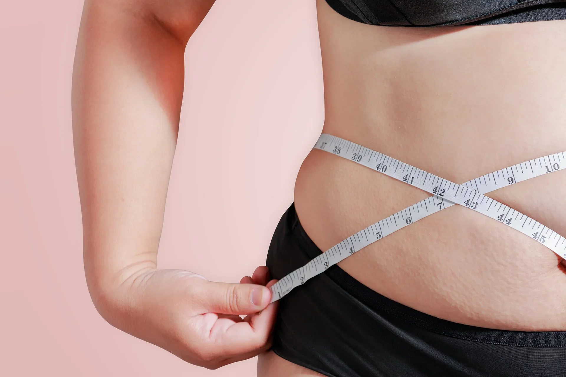 A person measuring their waist with a tape measure, focusing on midsection sculpting. They are partially dressed in black undergarments against a plain, light pink background.