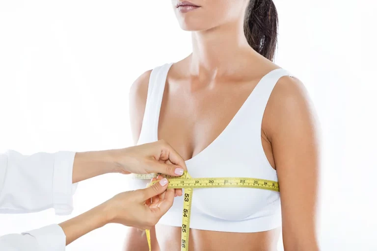 A person in a white bra is being measured around the bust with a yellow measuring tape held by another person, showcasing a step in natural breast augmentation.