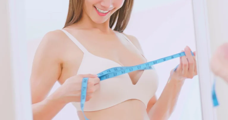 A woman in a light-colored bra measures her chest with a blue measuring tape, smiling at her reflection in the mirror, as if drafting the perfect fit.