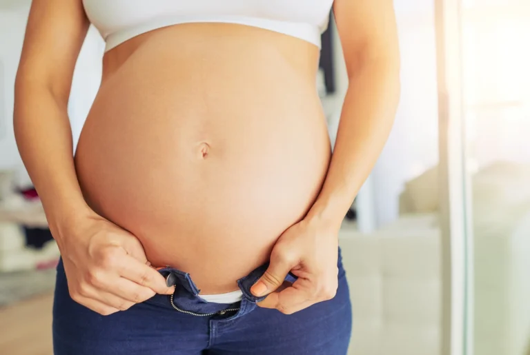 A pregnant person in a white top and blue jeans is seen safely buttoning their jeans.