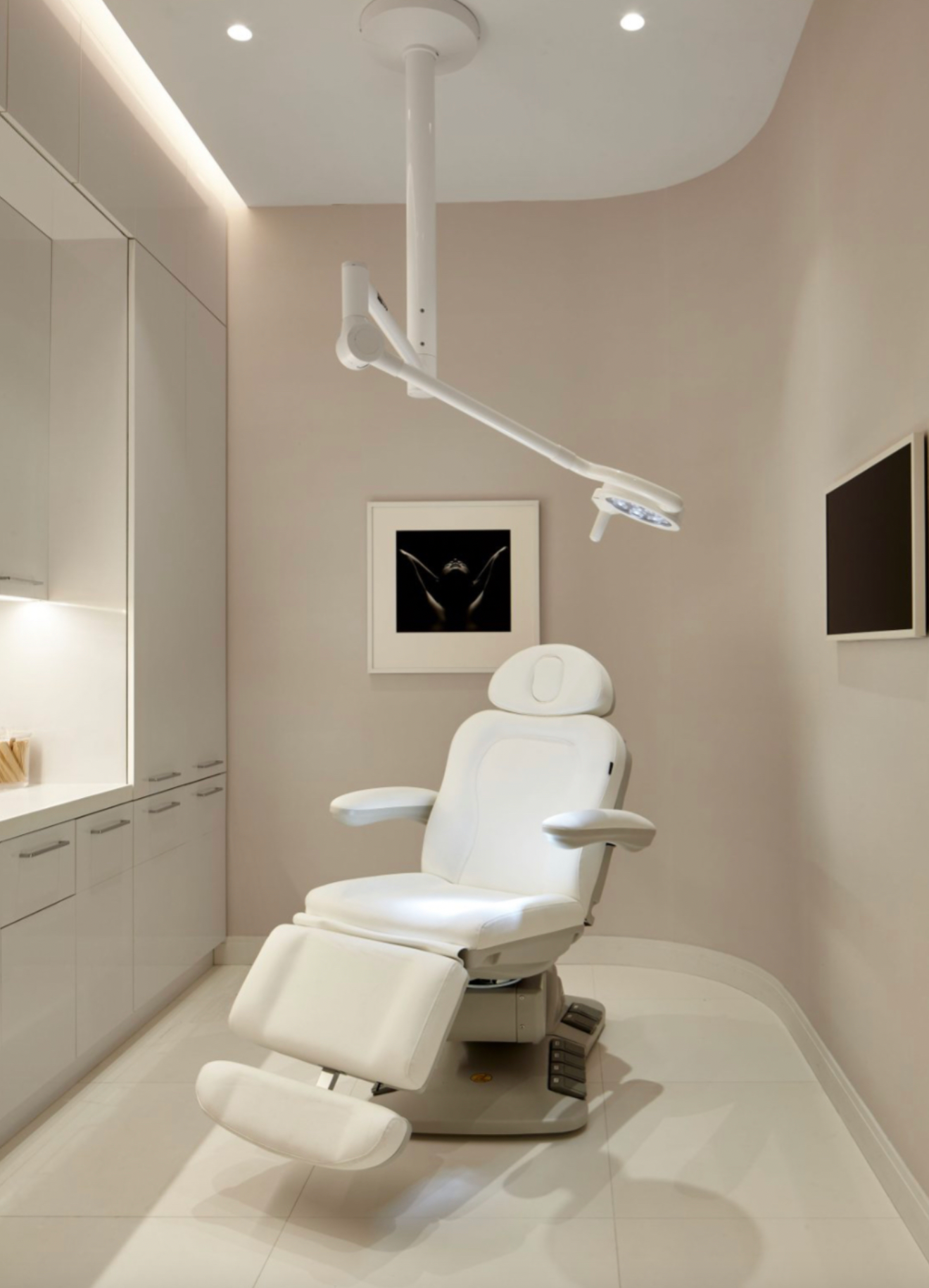 A modern, white medical examination room features a reclining chair, overhead light, cabinets, and framed artwork that subtly echoes the complexity of arms design.